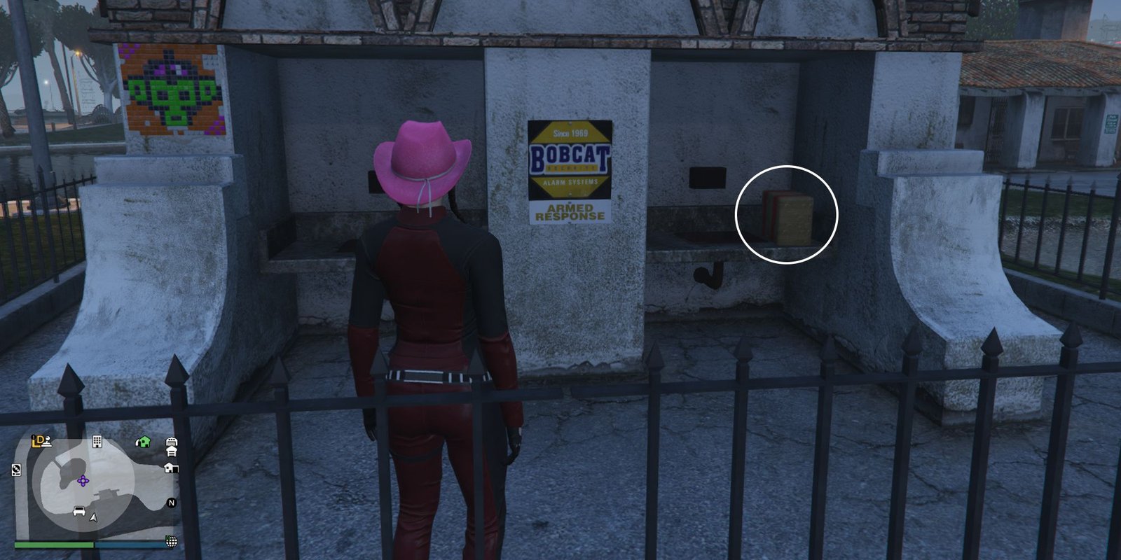 Image depicts a female player in GTA Online at the Mirror Park Lake stood opposite two sinks on the back of a toilet block, with a yellow sign that reads 'Bobcat' in the centre. On the right-hand sink, G's Cache is sat on the wall next to it.