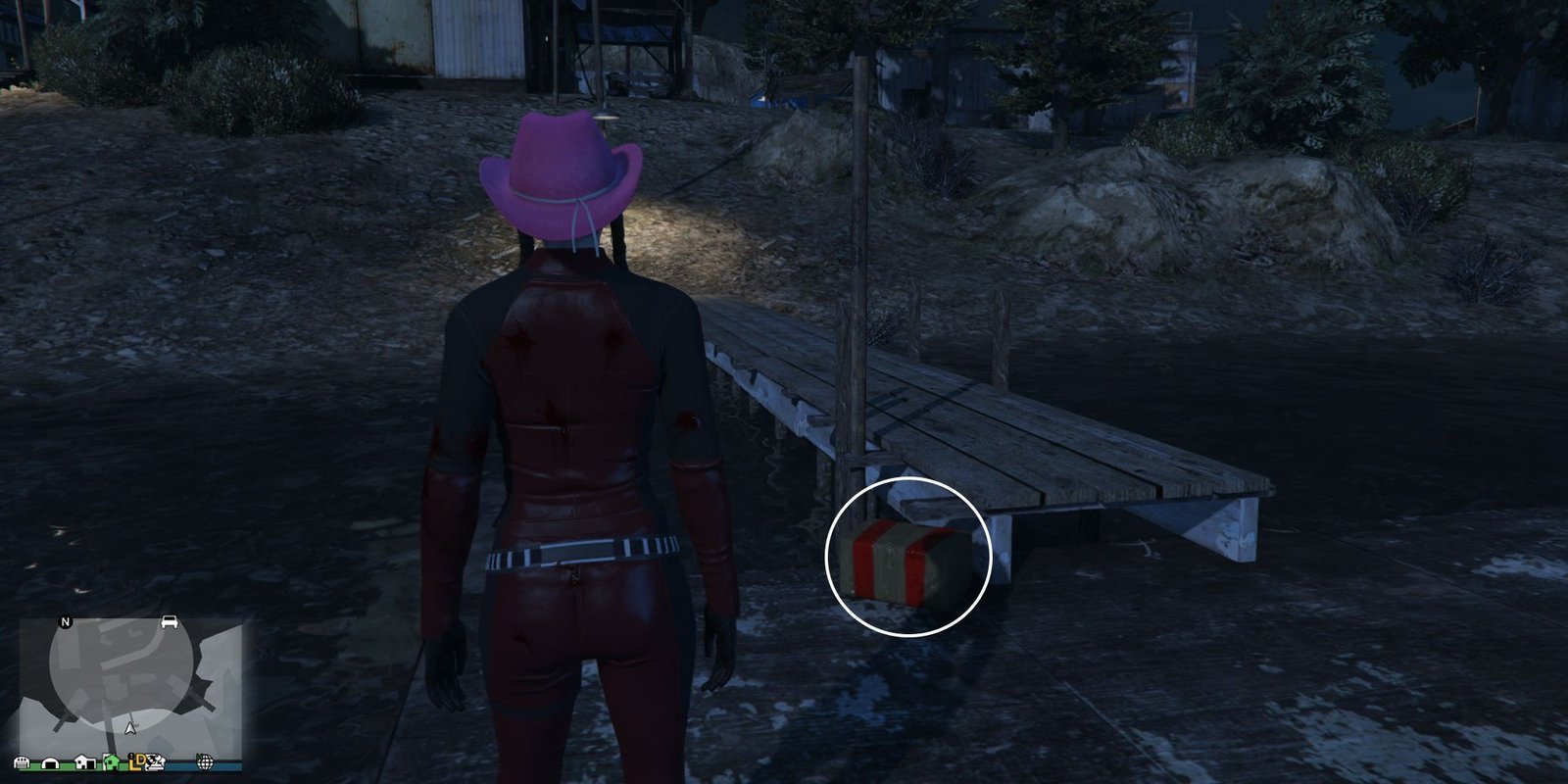 Image depicts a female character in GTA Online at Millar's Fishery stood on a platform at the end of a small dock on the water, facing towards land. At the end of the dock, the box for G's Cache is on the decking to the right of the player.