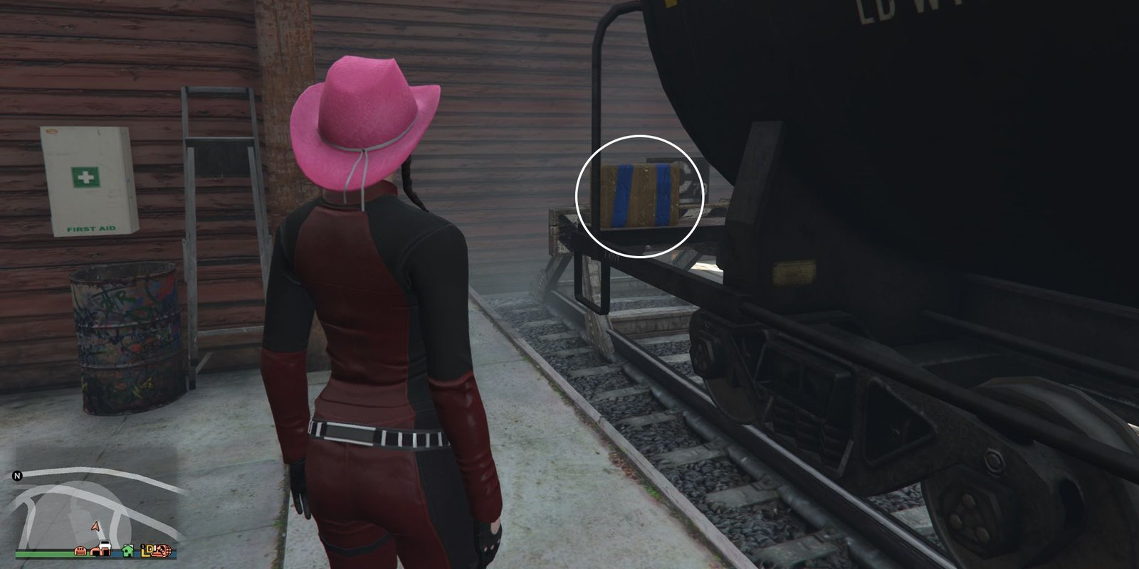 Image depicts a female character in GTA Online at Grapeseed Rail Terminus inside the railway shed next to a black tanker on the tracks. To the left of the player is a first aid kit, barrel, and ladder - and to the right is the G's Cache box on top of the end of the tanker.