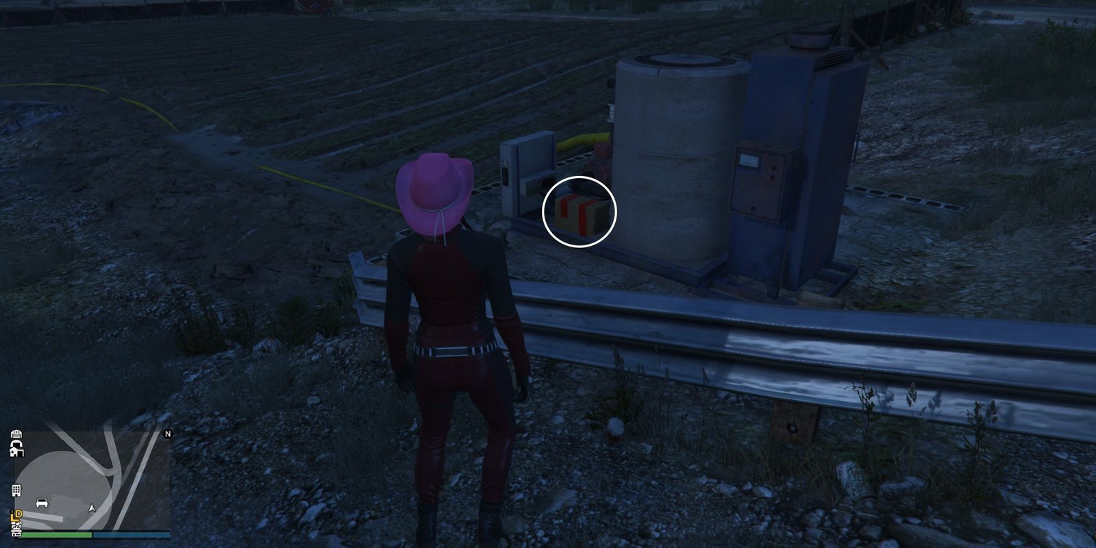 Image depicts a female character in GTA Online at Grapeseed Rail Terminus stood next to a water supply system overlooking some farming fields. Next to some of the water pipes is G's Cache.