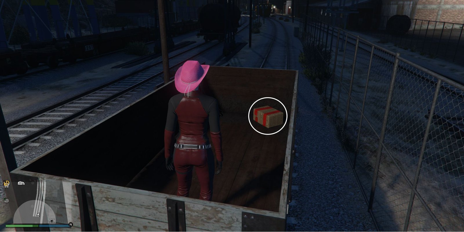 Image depicts a female character in GTA Online at Railyard Clucking Farms stood inside a wooden trailer on the railway tracks. In the back-right corner of this trailer is the G's Cache box.