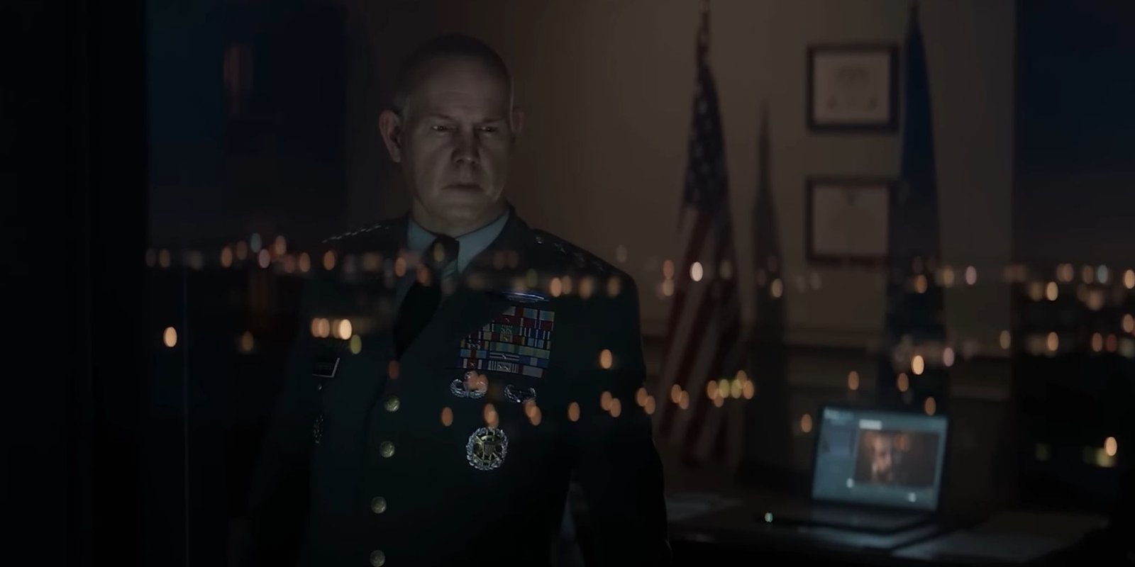 Shepherd turning his back away from Price and facing the glass window of his office, with the reflection of lights and the city skyline and Price on the laptop screen in the background.